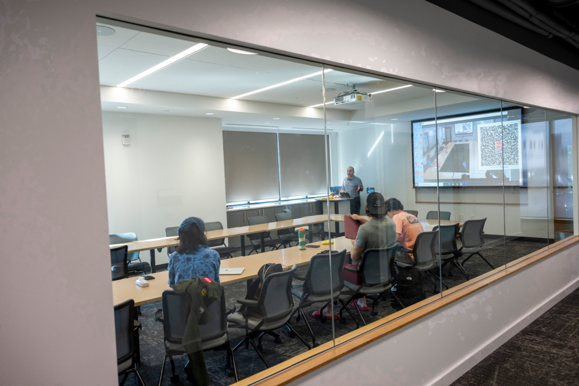 Severeal students sit in a classroom with a wall of windows, listening to a teacher.